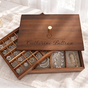 May include: A dark brown wooden jewelry box with a gold knob on the lid, engraved with the name Catherine Beltian and a heart symbol. The box features a removable top tray with multiple square compartments for rings and a larger section for necklaces.
