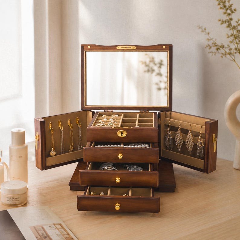 May include: A dark wood jewellery box featuring a hinged lid with an interior mirror, two side panels with gold-toned hooks for hanging necklaces, and three stacked drawers with gold-toned knobs for storing rings and other small accessories.