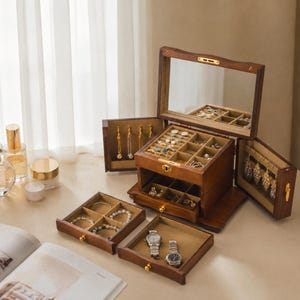 May include: A dark wood jewellery box with a hinged lid featuring an interior mirror, multiple open compartments, and side panels with hooks for hanging necklaces. The box includes two removable drawers with dividers for storing rings, bracelets, and two wristwatches.