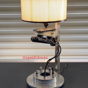 May include: A table lamp made from repurposed car parts. The lamp features a metal shade with a grid pattern, a brake master cylinder, and a brake rotor as the base. The lamp is lit and the shade is casting a warm glow. The lamp is signed "@speedlamps".