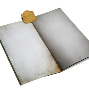 Può includere: Un libro aperto con pagine bianche sporche invecchiate e un segnalibro in metallo color oro. Le pagine del libro hanno un aspetto strutturato e i bordi mostrano una leggera scolorimento marrone. Il segnalibro è rettangolare con un design in rilievo.