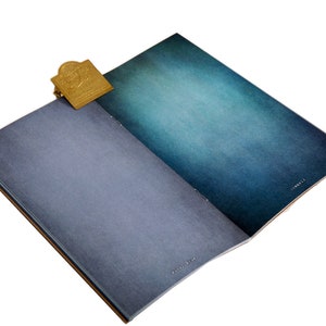 May include: An open notebook with blue and teal gradient pages. A gold-colored metal bookmark is attached to the top edge. The notebook has a textured cover and a brown spine.