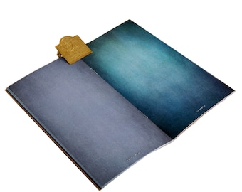 Puede incluir: Un cuaderno abierto con páginas degradadas azules y turquesas. Un marcapáginas de metal dorado está sujeto al borde superior. El cuaderno tiene una cubierta texturizada y un lomo marrón.