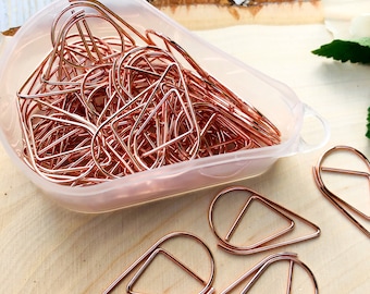 May include: A clear plastic container filled with rose gold paper clips shaped like teardrops. Several teardrop-shaped paper clips are scattered on a wooden surface.