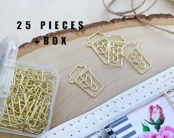 May include: A clear plastic box filled with 25 gold metal paper clips shaped like coffee cups with hearts. The box is sitting on a wooden surface with a piece of twine and a few of the clips scattered around it. The text "25 PIECES + BOX" is printed on the wooden surface.