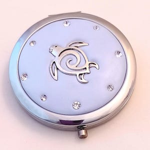 May include: A round, silver compact mirror with a light blue center. A silver sea turtle design is in the center, surrounded by clear rhinestones. The compact has a hinged opening and a small clasp.