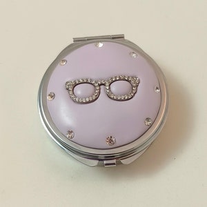 May include: A silver compact mirror with a lavender lid. The lid has a rhinestone-encrusted pair of glasses design.
