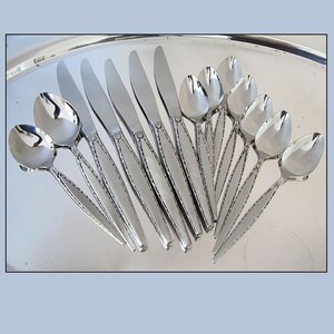 May include: A set of stainless steel flatware featuring five dinner knives and seven spoons. Each piece has a polished finish and a textured handle design with a repeating pattern along the edges, arranged in a fan shape on a silver tray.