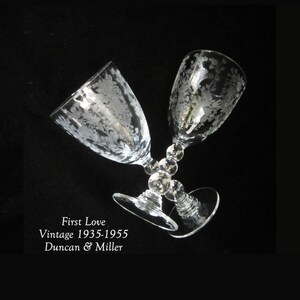 May include: Two clear glass wine goblets with a delicate etched floral pattern. The goblets are on a black background. The text "First Love Vintage 1935-1955 Duncan & Miller" is below the goblets.