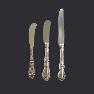 May include: Three silver butter knives with ornate, floral-patterned handles. The blades are curved and vary in size, with the largest knife on the right. The knives are set against a dark gray background.