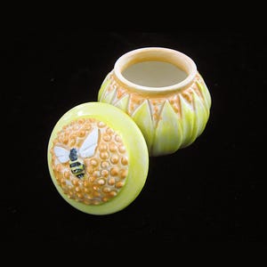 May include: A ceramic jar with a lid, featuring a bee and honeycomb design. The jar is yellow and orange, with a detailed bee illustration on the lid. The jar's body has a textured, petal-like design.