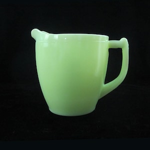 Puede incluir: Una jarra de crema verde claro de estilo vintage, con un asa curva y un pico vertedor. La jarra tiene un cuerpo redondeado y está hecha de vidrio translúcido verde pálido. El fondo es negro.