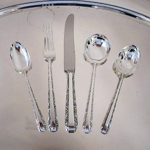 May include: A set of five silver-colored utensils: two spoons, a fork, a knife, and a serving spoon. The utensils have ornate, engraved handles and are arranged on a reflective silver tray.