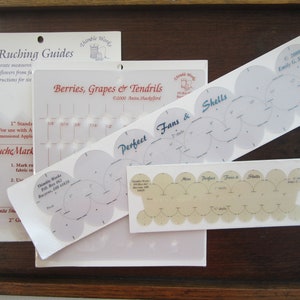 May include: Four white plastic templates for creating fabric ruffles. The templates are labeled "Strip Ruching Guides", "Berries, Grapes & Tendrils", "Perfect Fans & Shells", and "Fans & Shells".