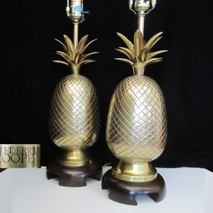 May include: A pair of gold pineapple table lamps with wooden bases. The lamps are made of brass and have a textured pineapple design. The lamps are in good condition and have a vintage look.