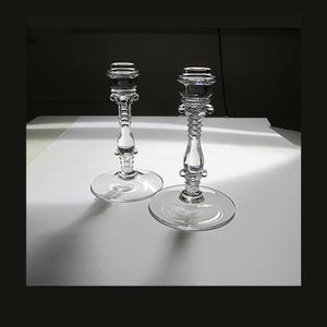 May include: A pair of clear glass candle holders featuring a tall, slender stem with decorative beaded detailing and a wide, circular base. The top of each holder has a flared rim designed to support a taper candle, with a smooth, transparent finish.