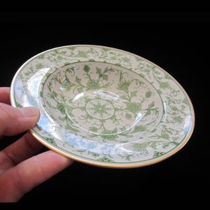 May include: A white ceramic bowl with a green floral design and a gold rim. The bowl is shallow and has a wide rim.