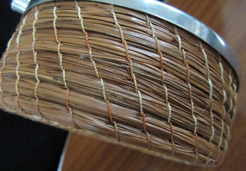 May include: A woven basket with a silver metal rim. The basket is made of natural brown fibres and has a woven pattern with white and orange threads.