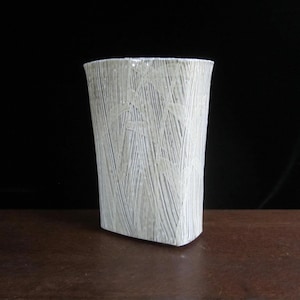 May include: A white ceramic vase with a textured, striped design. The vase has a rectangular shape and a slightly rounded base.