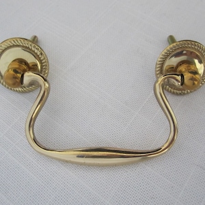 May include: A gold-toned drawer pull with a curved handle and two circular backplates. The backplates have a rope-like border. The handle is slightly curved and has a smooth, rounded design. The hardware is likely for furniture.