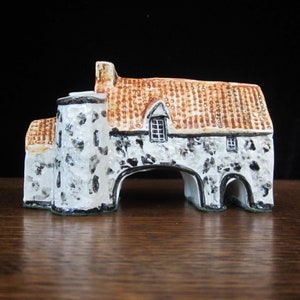 May include: A small, white and brown ceramic model of a house with a stone archway. The roof is brown and has a textured surface. The house has a small window and a chimney.