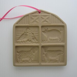 May include: A brown ceramic cookie cutter shaped like a barn with four squares. Each square has a different farm animal design: a pig, a cow, a sheep, and a rooster. The barn has a red ribbon attached to the top.
