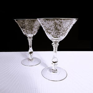 May include: Two clear, stemmed cocktail glasses with etched floral designs. The glasses have a wide, triangular bowl and a detailed stem. The base is a simple, round disc. The background is black and white.