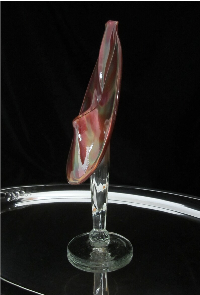 May include: A glass vase with a unique, abstract design. The vase is made of clear glass with a red, brown, and green iridescent finish. The vase has a tall, slender stem and a wide, open mouth.