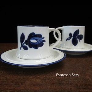 May include: Two white espresso cups with blue floral designs and matching saucers. The cups have a blue trim around the rim and the saucers have a blue band around the edge. Espresso Sets.