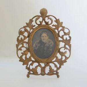 May include: Ornate gold-colored oval picture frame with a black and white portrait of a woman. The frame has a decorative, openwork design with a stand on the back. The portrait is centered within the frame.