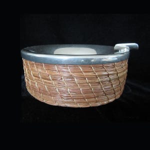 May include: A silver and woven wicker ashtray with a removable lid. The ashtray is round and has a diameter of approximately 15 cm.