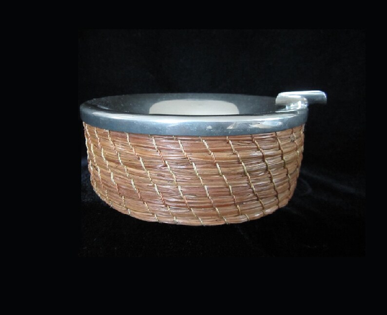 May include: A silver and woven wicker ashtray with a removable lid. The ashtray is round and has a diameter of approximately 15 cm.
