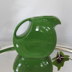 May include: A green ceramic pitcher with a unique, modern design. The pitcher has a wide, rounded base and a curved spout. It is made of a glossy, smooth ceramic.