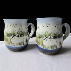May include: Two ceramic mugs with blue and white glaze. Each mug features a hand-painted design of a white dog with black spots running through tall grass with birds flying overhead.