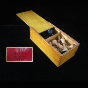 May include: A rectangular wooden box with a sliding lid containing light-colored wooden chess pieces. A separate red rectangular label is visible, featuring the text STAUNTON, along with other partially legible printed information.