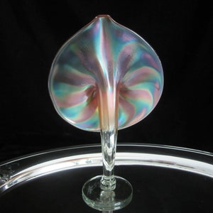 May include: A iridescent glass vase in the shape of a calla lily with a pink, blue, and green color scheme. The vase is sitting on a silver tray.
