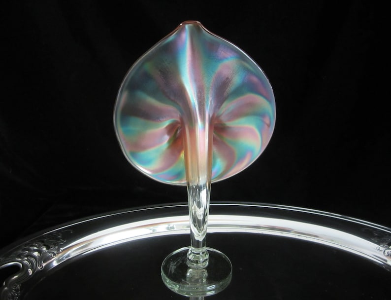 May include: A iridescent glass vase in the shape of a calla lily with a pink, blue, and green color scheme. The vase is sitting on a silver tray.