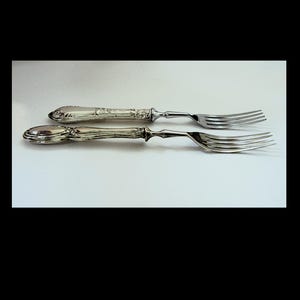 May include: Two silver-toned metal forks with ornate, textured handles and four tines each. The handles feature intricate embossed detailing and a rounded end, while the neck of each fork transitions into a smooth, polished finish for the tines.