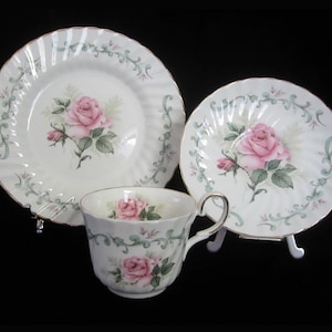 May include: A white teacup and saucer set with a pink rose design and green leafy accents. The set has a gold rim and is decorated with a delicate floral pattern.