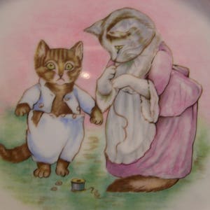 May include: A vintage porcelain plate with a hand-painted illustration of two kittens. One kitten is wearing a pink dress and the other is wearing a blue and white outfit. The plate has a pink background and a white border.