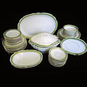 May include: A complete white porcelain dinnerware set with a green and gold trim. The set includes plates, bowls, saucers, and a serving dish with a lid. The pieces are arranged on a black surface.