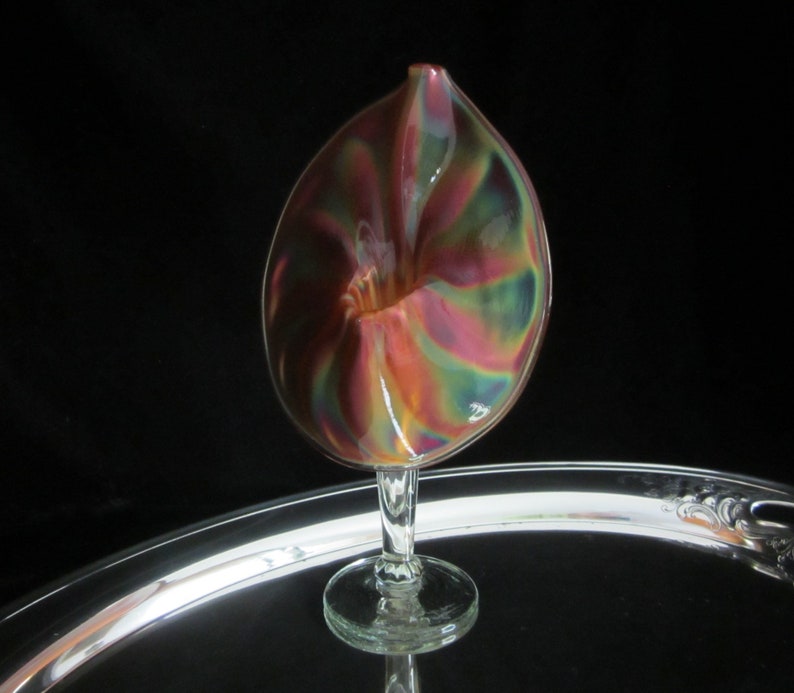 May include: A iridescent glass vase with a unique swirl design. The vase is shaped like a flower and has a clear glass stem.