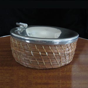 May include: A silver ashtray with a woven straw base. The ashtray has a small metal lip around the top and a metal tab on the side.