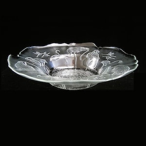 May include: Clear glass decorative bowl with a scalloped edge. The bowl features etched designs of birds, plants, and water. The bowl is round with a textured center and sits on a black background.