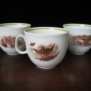 May include: Three white porcelain teacups with green rims and brown illustrations of birds and landscapes. The cups are on a dark wood surface.