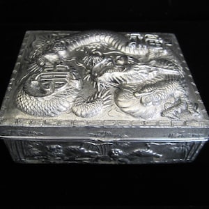 May include: A silver metal box with an intricate design of dragons and Chinese characters. The box is square and has a hinged lid.