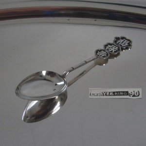 May include: A silver spoon with a rounded bowl and a long handle. The handle is decorated with three round, black and white Chinese characters. The spoon is lying on a silver tray.
