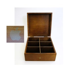 May include: A square, dark brown wooden box with an open hinged lid and six internal compartments. The front of the box and the lid feature a small, light-colored apple logo. The box has a smooth finish and visible wood grain throughout the structure.