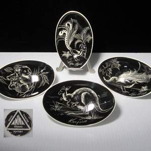 May include: Set of four black and white ceramic plates with Australian animal illustrations. Each plate features a different animal, including a koala, kangaroo, kookaburra, and lyrebird. The plates are oval shaped and have a white rim. The plates are marked with "Anna Studio Pottery".