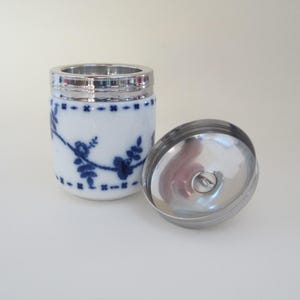 May include: A white ceramic jar with a blue floral design and a silver lid. The lid is open and the inside of the jar is visible.